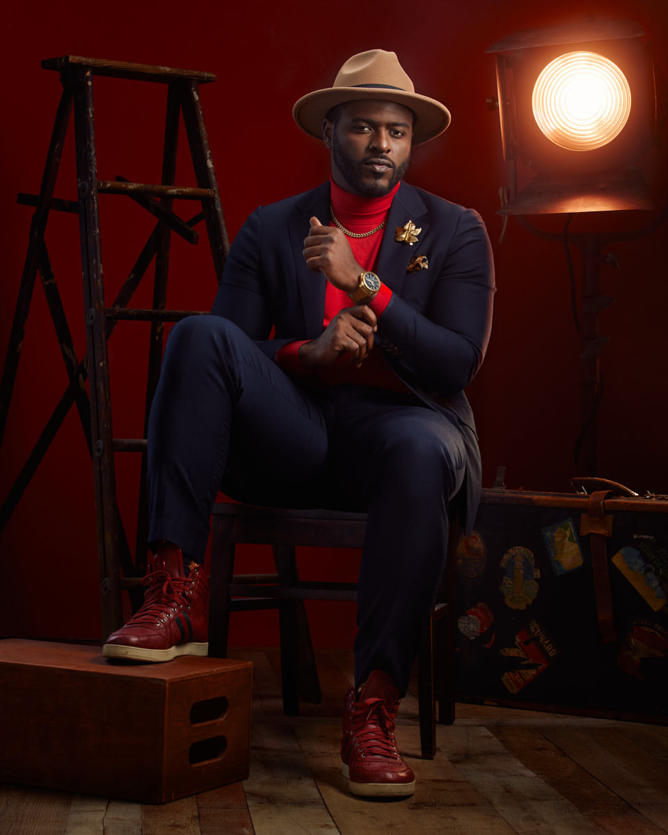 Man in a suit and hat sitting on a stool with a spotlight in a dramatic setting